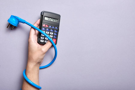 Conceptual image about the high economic cost of electric power and the increase in energy rates: power cable trapping an arm, the hand holds a calculator.の写真素材