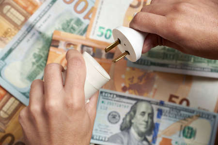 Hands plugging or unplugging a socket against a background of dollar and euro bills. Concepts of...