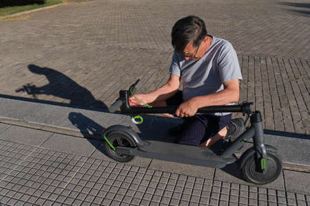 Hispanic senior man unfolding his electric kick scooter in a city park on a sunny morning. Concepts...