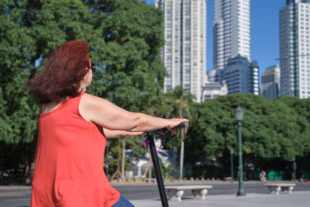 Hispanic mature woman riding her electric kick scooter around the city in summer. Concepts of sustainability, summer tourism, active life, clean energy and green mobility, eco transportation.の写真素材