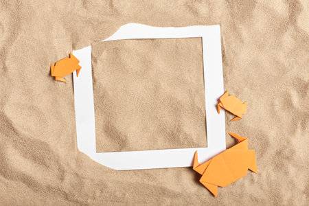 Fun top view composition, concepts of summer, beach and vacations: smooth sand background with a family of origami crabs and an empty white paper frame. Creative layout, minimal design with copyspace.の写真素材