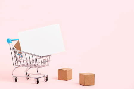 Shopping cart with cardboard boxes and a blank white paper poster on a pastel pink background. Minimal design with copy space. Concepts: market deals, black friday, logistics and packaging.の写真素材