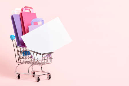 Shopping cart loaded with paper bags and a blank card on a pastel pink background. Minimal design with copy space. Concepts: market deals, seasonal sales and discounts, black friday.の写真素材