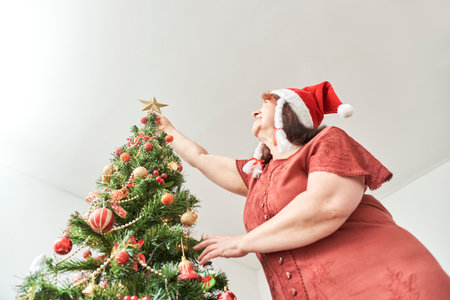Mature latin woman putting a gold star on the top of her Christmas tree wearing a red Santa Claus beanie. The joy of spending the holidays at home.の写真素材