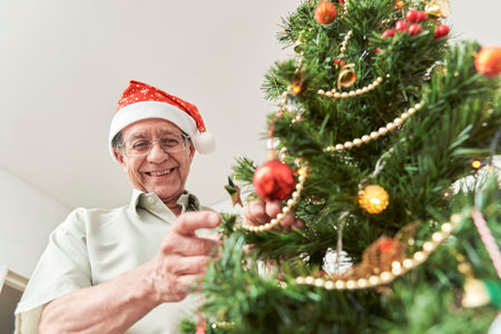 Happy senior hispanic man smiling while decorating a Christmas tree at home wearing a red Santa Claus beanie. The happiness of the holidays. Low angle view with copy space.の写真素材