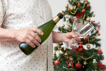 Unrecognizable man holding a bottle of champagne or cider and a pair of glasses with a Christmas tree in the background. Concept of celebratory toast.の写真素材