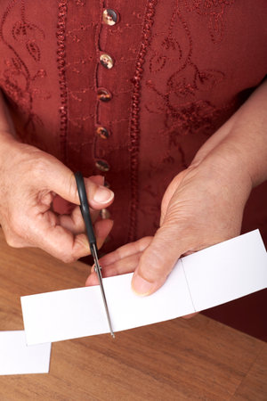 Unrecognizable mature woman cutting with scissors white cardboard, making handmade cards to put on...