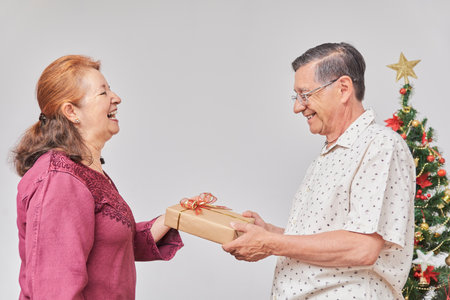 Senior Hispanic couple happily spending the holidays at home, one hands the other a Christmas gift...