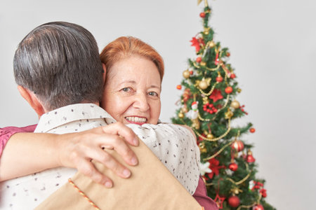 Senior latin couple celebrating the holidays together at home, they happily embrace as they exchange Christmas presents, she looks at the camera while holding her giftbox.の写真素材