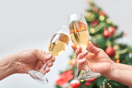 Unknown adult couple toasting during Christmas or New Year, celebrating holidays together. Low angle view of a female and male hand clinking a pair of glasses, with a decorated tree in the backgroundの写真素材