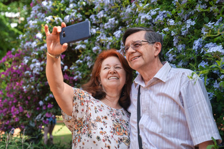 Happy senior hispanic couple taking a self portrait with a smartphone in a garden, surrounded by...
