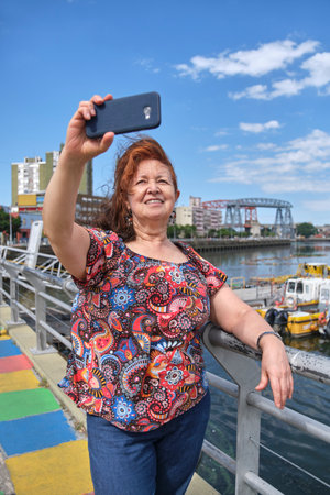 Mature latin woman taking a selfie with her smartphone in the touristic neighborhood of La Boca, in Buenos Aires, Argentina, on a sunny summer day.の写真素材