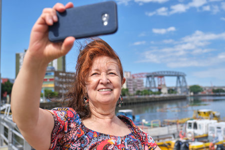 Mature latin woman taking a selfie with her smartphone in the touristic neighborhood of La Boca, in Buenos Aires, Argentina, on a sunny summer day.の写真素材