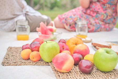 Unrecognizable couple having a picnic breakfast with a variety of fresh fruits, apples, peaches, tangerines, plums, watermelon. Focus on them. Concepts: healthy eating, enjoying the outdoors at summerの写真素材