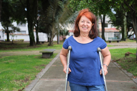 Mature hispanic human recovering from an injury standing with crutches on an urban park pathway, smiling. Concepts: positive attitude towards adversity, physical rehabilitationの写真素材