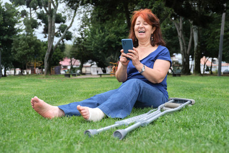 Mature hispanic woman resting on the grass, laughing while using her smartphone, barefoot, with a bandage on her left foot and crutches at her side. Concepts: positive attitude towards an injury.の写真素材