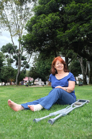 Mature woman resting on the grass, smiling, barefoot, with a bandage on her left foot and crutches at her side. Concepts: positive attitude towards an injury, recovery and physical rehabilitation.の写真素材