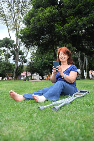Mature hispanic woman resting on the grass, smiling while using her smartphone, barefoot, with a bandage on her left foot and crutches at her side. Concepts: positive attitude towards an injury.の写真素材