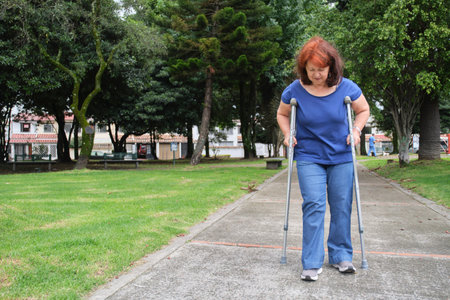 Mature hispanic human recovering from an injury walking with crutches on an urban park pathway, smiling. Concepts: positive attitude towards adversity, physical rehabilitationの写真素材