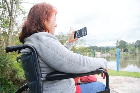 Latin mature woman in wheelchair smiling while talking via video call from her smartphone outdoors....