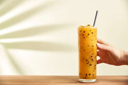 Hand gripping a glass of a refreshing passion fruit drink, juice or cocktail, with a reusable ecological straw on a wooden surface. The shadow of a tropical leaf is projected on the back wall.の写真素材