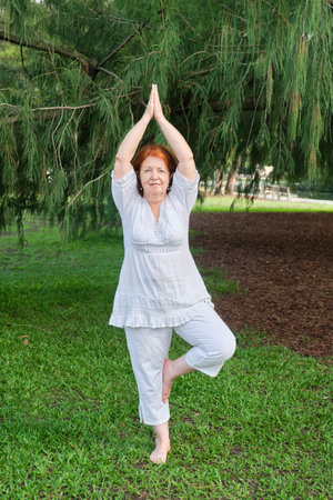 Mature Hispanic woman in white clothes practicing yoga outdoors in a park. Vrksasana standing pose. Concepts: wellness, vitality, active and healthy lifestyle.の写真素材