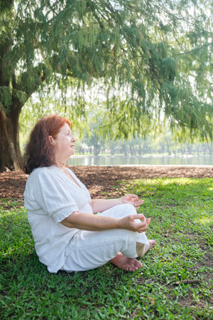 Portrait of a mature Latin woman in white clothes sitting practicing yoga and meditation outdoors in a park. Concepts: wellness, tranquility and mental health.の写真素材