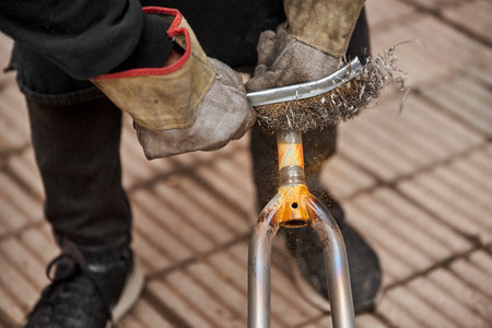 Unrecognizable man with protective gloves using a wire brush over a bicycle fork after passing a blowtorch over it to remove the paint, as part of his bike renovation work. Close up view.の写真素材