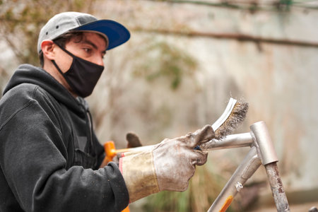Young Hispanic man removing the paint from an orange bicycle frame with a wire brush, as part of a bike renovation work made at his workshop. Selective focus composition with copy space.の写真素材