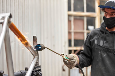 Young Hispanic man using a blowtorch to remove the paint from an orange bicycle frame that is being renovated in his workshop.の写真素材