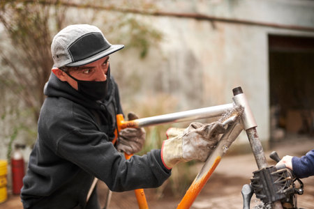 Young Hispanic man removing the paint from an orange bicycle frame, as part of a bike renovation work made at his workshop.の写真素材