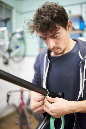 Hispanic young man preparing a bicycle frame with masking tape for a custom painting design in his...
