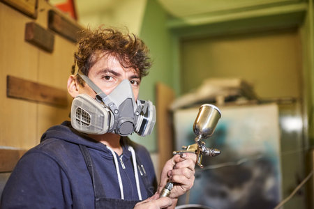 Portrait of a latin young worker wearing a protective respirator mask looking at camera holding an airbrush during the process of spray painting a bicycle in his workshop. Real people at work.の写真素材