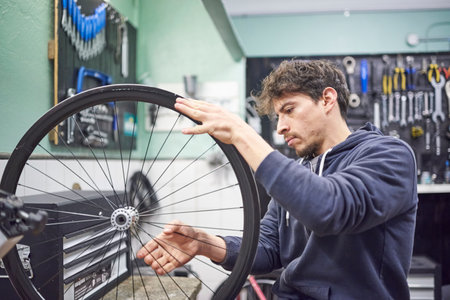 Young hispanic man checking the tension of the spokes of a bicycle wheel in his bike workshop. Real people at work.の写真素材
