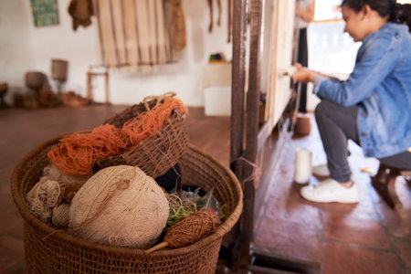 Barichara, Santander, Colombia; November 25, 2022: Basket with fibers and balls of natural materials for weaving. In the background a young woman learning the traditional art of loom weaving.のeditorial素材