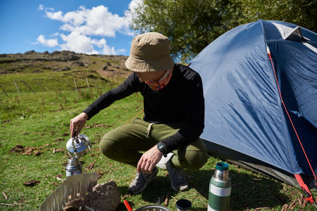 Hispanic young man pouring hot water from a kettle into a thermos, outdoors, at the side of his tent...
