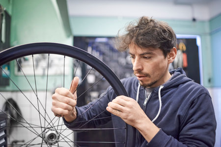 Young hispanic man checking the tension of the spokes of a bicycle wheel in his bike workshop. Real people at work.の写真素材
