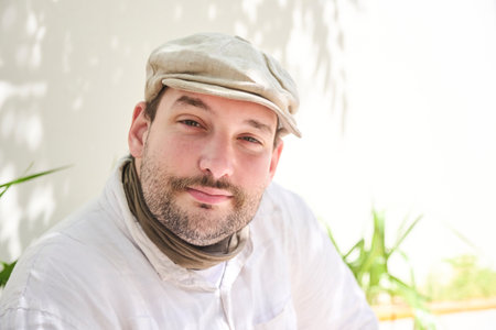 Close up portrait of a young man sitting in a sunny courtyard, looking at the camera while smiling....