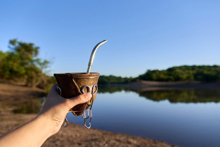 An unrecognizable person holds out a hand with a mate, in the background is an Argentinian landscape, the bank of a quiet stream in the morning sun.の写真素材