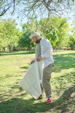 Young man outdoors choosing a suitable place to set up his camp. He is holding in his hands the plastic that will go under the tent. The environment is green and the weather is sunny.の写真素材