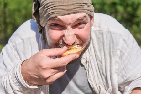 Playful portrait of a young man outdoors looking at the camera while eagerly eating his breakfast,...