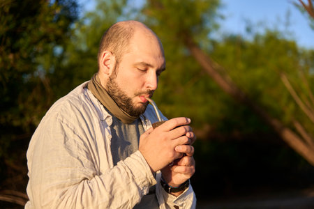 Hispanic male traveler sitting on the bank of a stream at dawn, receiving the first rays of sunshine as he drinks mate, a typical Argentine drink. National Park, Entre Rios, Argentina.の写真素材