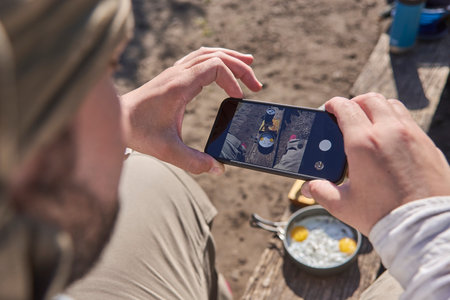 Man photographing a camping breakfast, fried eggs and toasted bread, with a phone to share it on social media. Concept: technology makes it possible to be traveling in the wild but still be connectedの写真素材