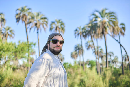 Portrait of a young man with sunglasses walking outdoors on a sunny day, with palm trees in the...