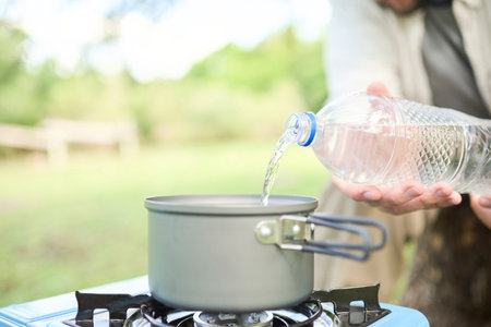 Unrecognizable person pouring drinking water into a pot as part of the process of cooking outdoors, in a portable gas stove, while camping. Selective focus on water. Close up view, with copy space.の写真素材