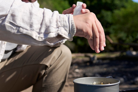 Unrecognizable male traveler seasoning his food, cooking outdoors while camping at a nature campground a sunny morning. Close up view of the hand that pours the salt.の写真素材
