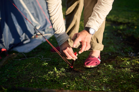 Unrecognizable person using a stone as a hammer to drive a tent peg into the ground, from the tent he is setting up in a camp, at sunset.の写真素材