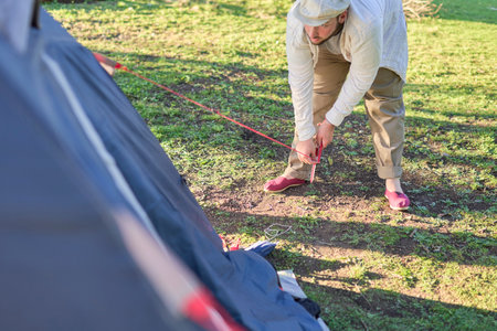 Young male traveler adjusts the tension of his tent rainfly while setting up camp in the nature at dusk.の写真素材