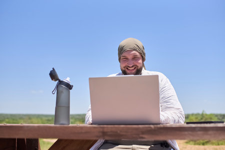 Young Hispanic man smiling while working on his laptop at an outdoor table on a sunny day. Concept: digital nomad working remotely while traveling enjoying a lifestyle of freedom.の写真素材