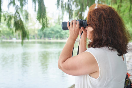 Senior Hispanic woman looking into the distance, using binoculars to watch birds as a hobby in an urban park with trees and a lake. Composition with copy space.の写真素材